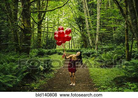 Stock Photo - Korean woman holding red balloons in lush forest. Fotosearch