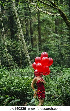 Stock Photograph - Korean woman holding red balloons in lush forest. Fotosearch