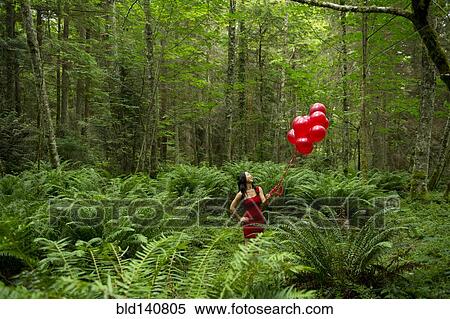 Stock Photography - Korean woman holding red balloons in lush forest. Fotosearch