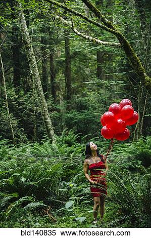 Stock Photography - Korean woman holding red balloons in lush forest. Fotosearch