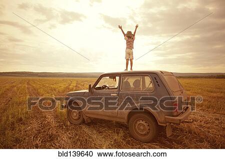 Stock Image - Mari boy standing on car roof in rural field. Fotosearch