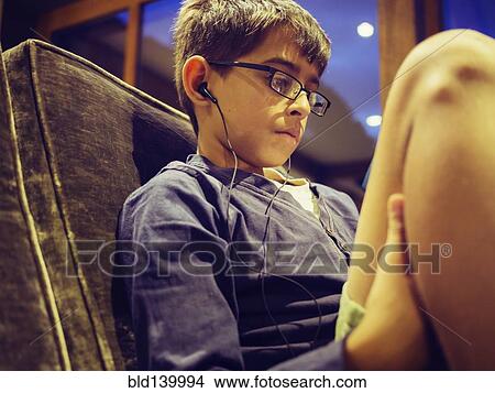 Picture - Mixed race boy listening to earbuds on sofa. Fotosearch