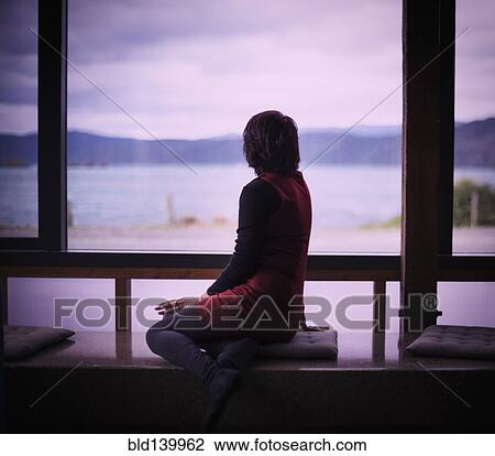 Stock Image - Mixed race woman looking out window of modern house. Fotosearch