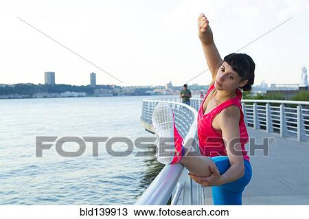 Stock Image - Mixed race woman stretching on urban waterfront railing, . Fotosearch