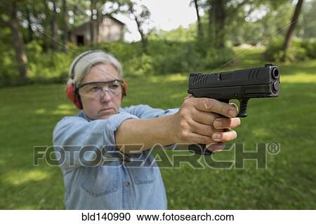 Stock Image - Older Caucasian woman practicing with gun at shooting range. Fotosearch