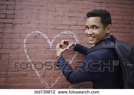 Stock Image - Smiling man making heart shape with hands by heart chalk drawing on wall. Fotosearch