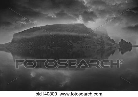 Stock Image - Snowy mountain under cloudy sky reflected in lake. Fotosearch