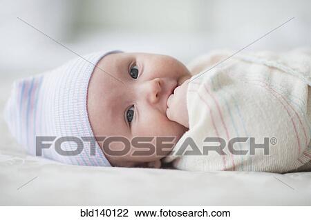 Stock Image - Swaddled Asian baby biting fingers on bed. Fotosearch