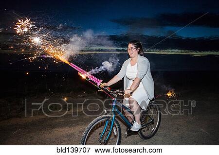 Stock Photo - Woman holding fireworks on bicycle at night. Fotosearch