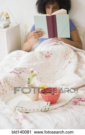Stock Image - Woman reading book and with breakfast in bed. Fotosearch