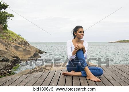 Stock Image - Woman sitting on wooden deck overlooking lake. Fotosearch