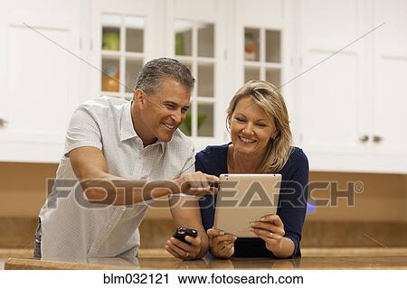 Stock Image - Caucasian couple using digital tablet and cell phone at kitchen counter. Fotosearch