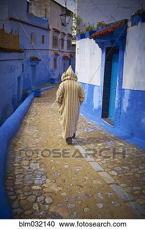 Stock Image - Caucasian hooded man walking on colorful historical village street, Chefchaouen, Chefchaouen, Morocco. Fotosearch