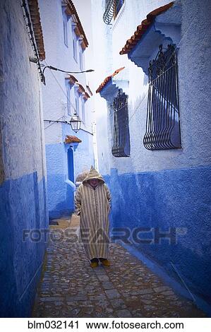 Stock Image - Caucasian hooded man walking on colorful historical village street, Chefchaouen, Chefchaouen, Morocco. Fotosearch