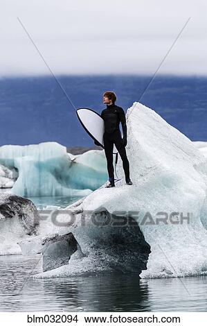 Picture - Caucasian surfer carrying board near glacial water. Fotosearch