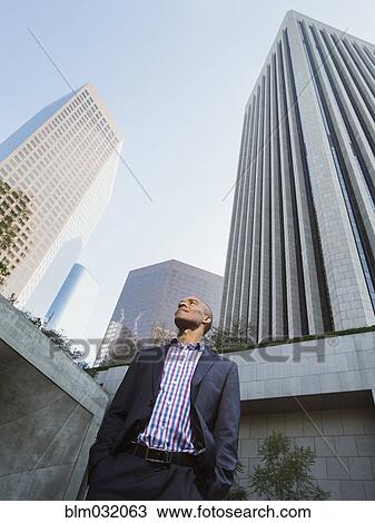 Stock Image - Low angle view of Black businessman standing outside urban skyscraper. Fotosearch