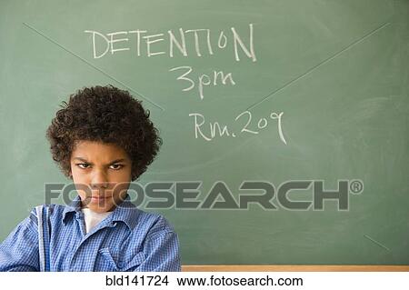 Picture - Angry mixed race boy frowning in classroom detention. Fotosearch