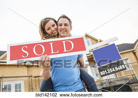 Stock Image - Caucasian couple holding sold sign near house under construction. Fotosearch