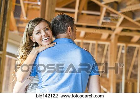 Stock Image - Caucasian couple hugging in house under construction. Fotosearch