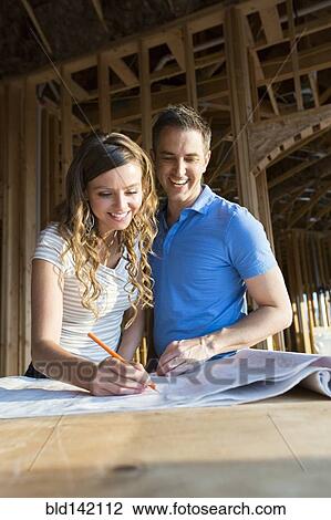 Stock Image - Caucasian couple marking blueprints in house under construction. Fotosearch