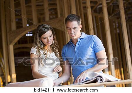 Stock Image - Caucasian couple reading blueprints in house under construction. Fotosearch