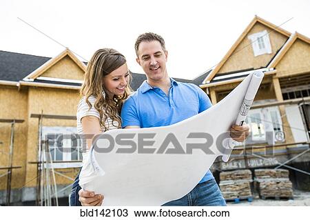 Stock Image - Caucasian couple reading blueprints outside house under construction. Fotosearch