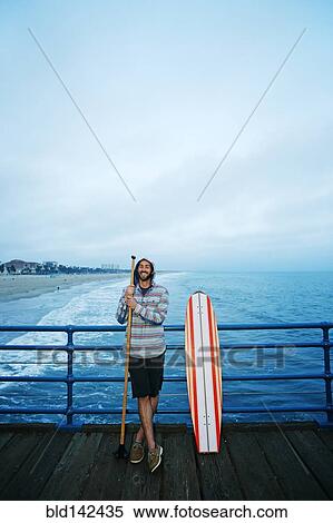Stock Photography - Caucasian man standing with skateboard and paddle pole on boardwalk. Fotosearch