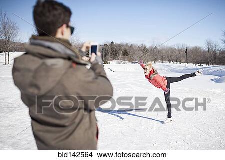 Picture - Caucasian man taking picture of girlfriend ice skating on frozen lake. Fotosearch