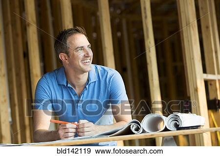 Stock Photo - Caucasian man with blueprints in house under construction. Fotosearch