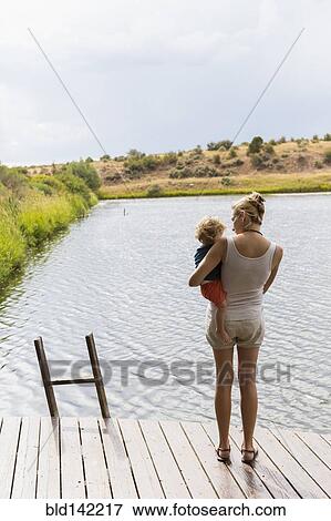 Stock Photo - Caucasian mother holding baby son on wooden deck near lake. Fotosearch