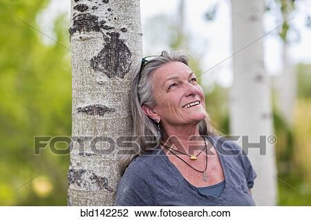 Stock Image - Caucasian senior woman leaning on tree in forest. Fotosearch