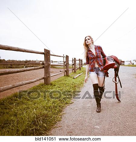 Stock Image - Caucasian woman carrying horse saddle on ranch. Fotosearch