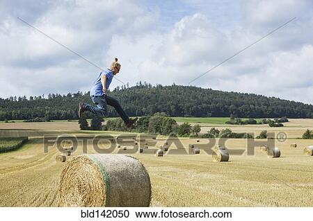 Stock Image - Farmer jumping over hay bale in field. Fotosearch