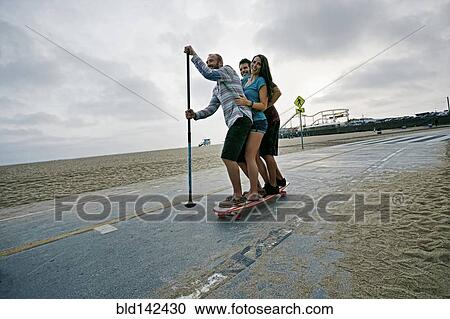 Stock Image - Friends skateboarding with paddle pole at beach. Fotosearch