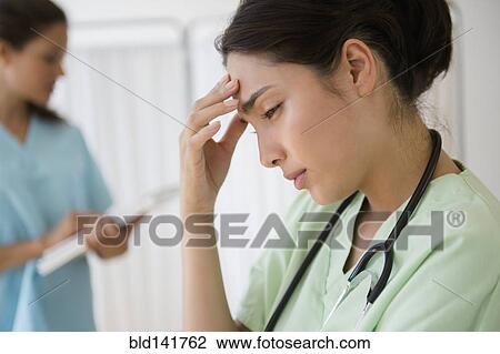 Stock Image - Hispanic nurse rubbing forehead at hospital. Fotosearch