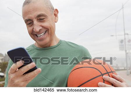 Stock Photography - Hispanic senior man with basketball using cell phone. Fotosearch
