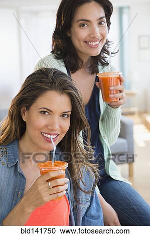 Stock Image - Hispanic women drinking healthy carrot juice in living room. Fotosearch