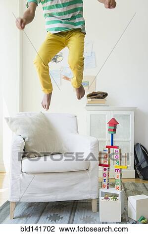 Stock Image - Mixed race boy jumping on armchair in living room. Fotosearch