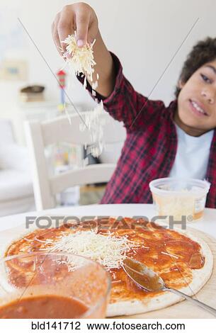 Stock Image - Mixed race boy sprinkling cheese on homemade pizza. Fotosearch