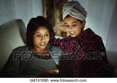 Stock Image - Mixed race mother and daughter using digital tablet at night. Fotosearch