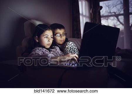 Stock Image - Mixed race sisters using laptop at night. Fotosearch