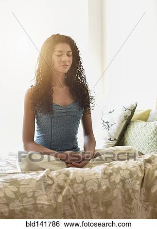 Stock Photograph - Mixed race woman meditating on bed. Fotosearch