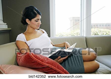 Stock Image - Mixed race woman reading book on sofa. Fotosearch