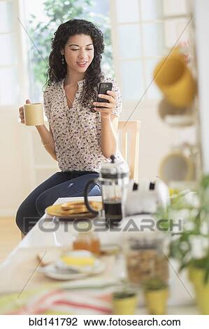 Stock Image - Mixed race woman using cell phone in kitchen. Fotosearch