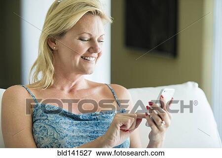 Stock Photo - Smiling woman using cell phone on sofa. Fotosearch