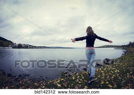 Stock Image - Woman standing with arms outstretched near lake. Fotosearch