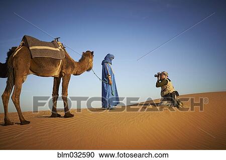 Stock Image - Tourist photographing guide with camel on sand dune in desert landscape. Fotosearch