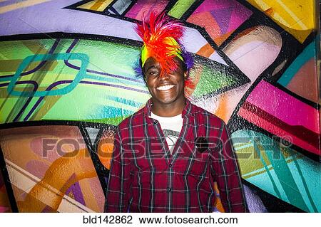 Stock Image - Black man in colorful wig smiling near graffiti wall. Fotosearch