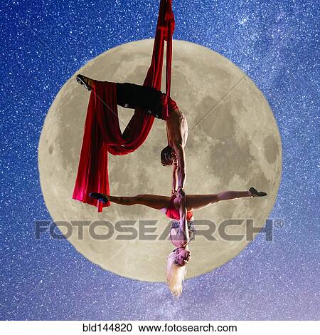 Stock Image - Caucasian acrobats performing with silk ropes by full moon. Fotosearch