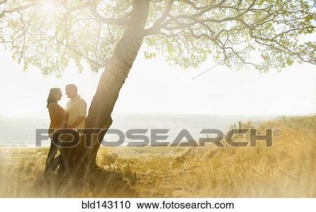 Stock Image - Caucasian couple hugging near tree in field. Fotosearch